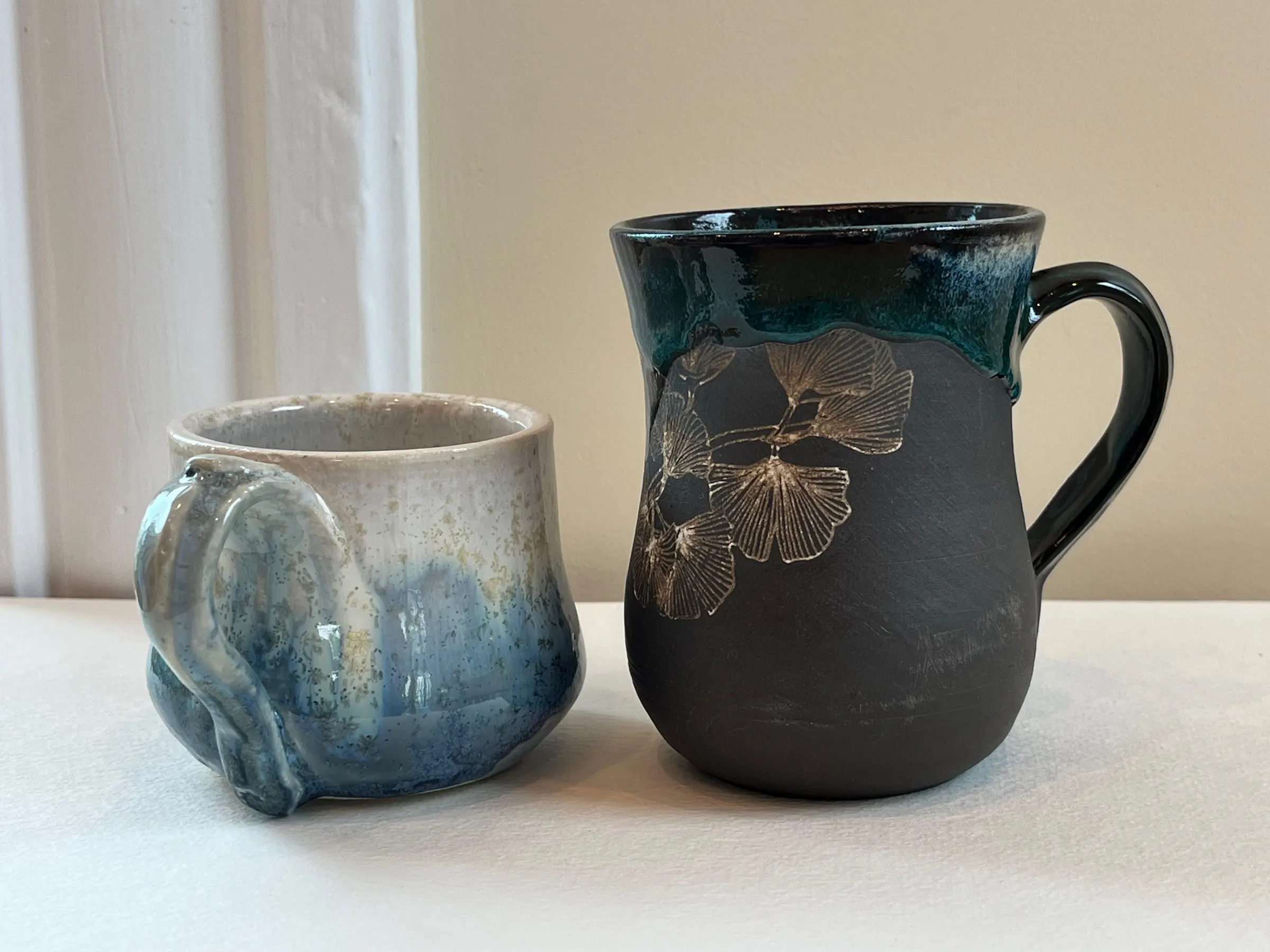 A black mug with a carved ginkgo leaf and teal drip beside a blue and cream mug with a hippo-shaped handle