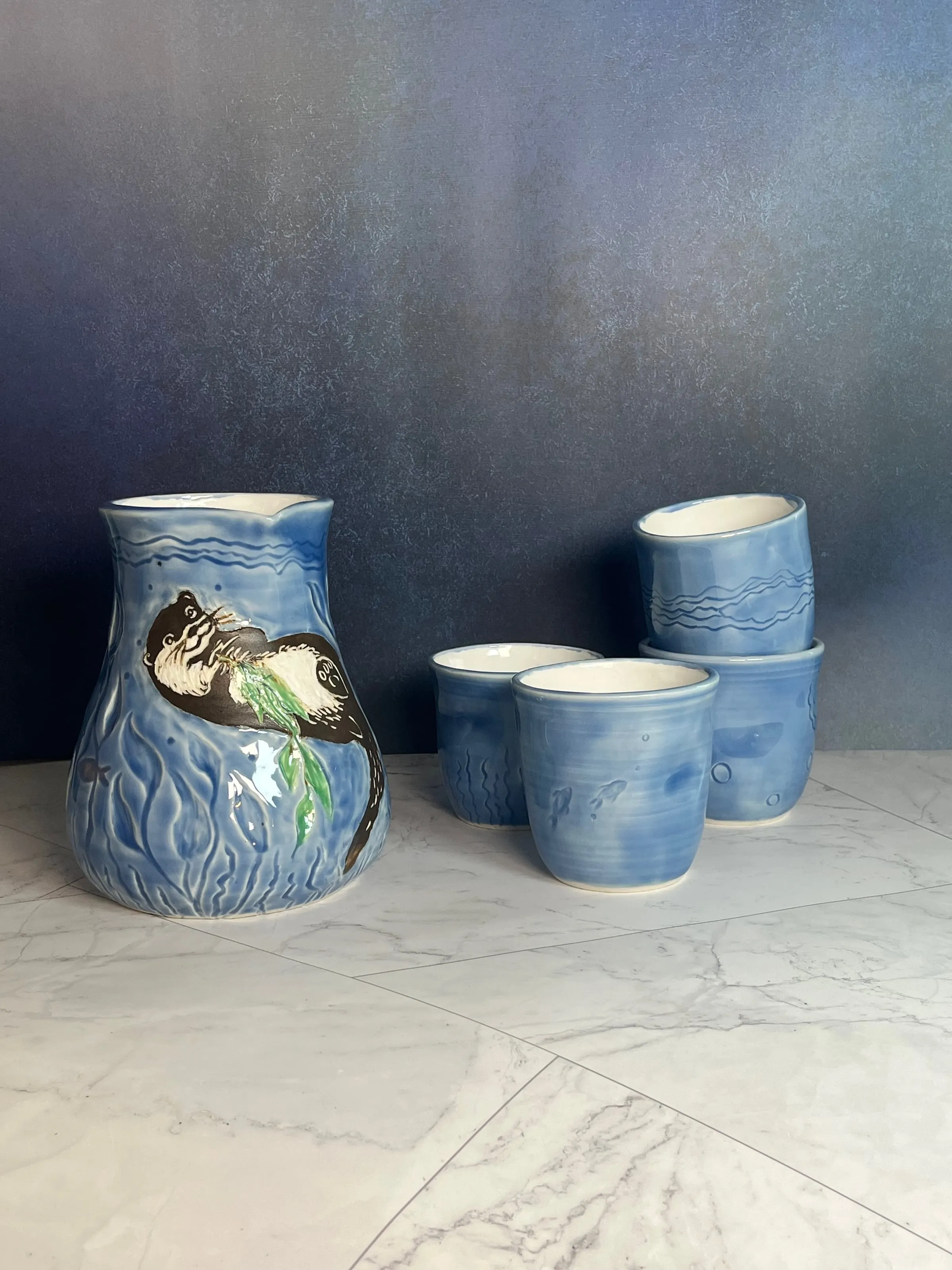 A blue stoneware pitcher with a carved otter holding seaweed, beside four matching blue cups