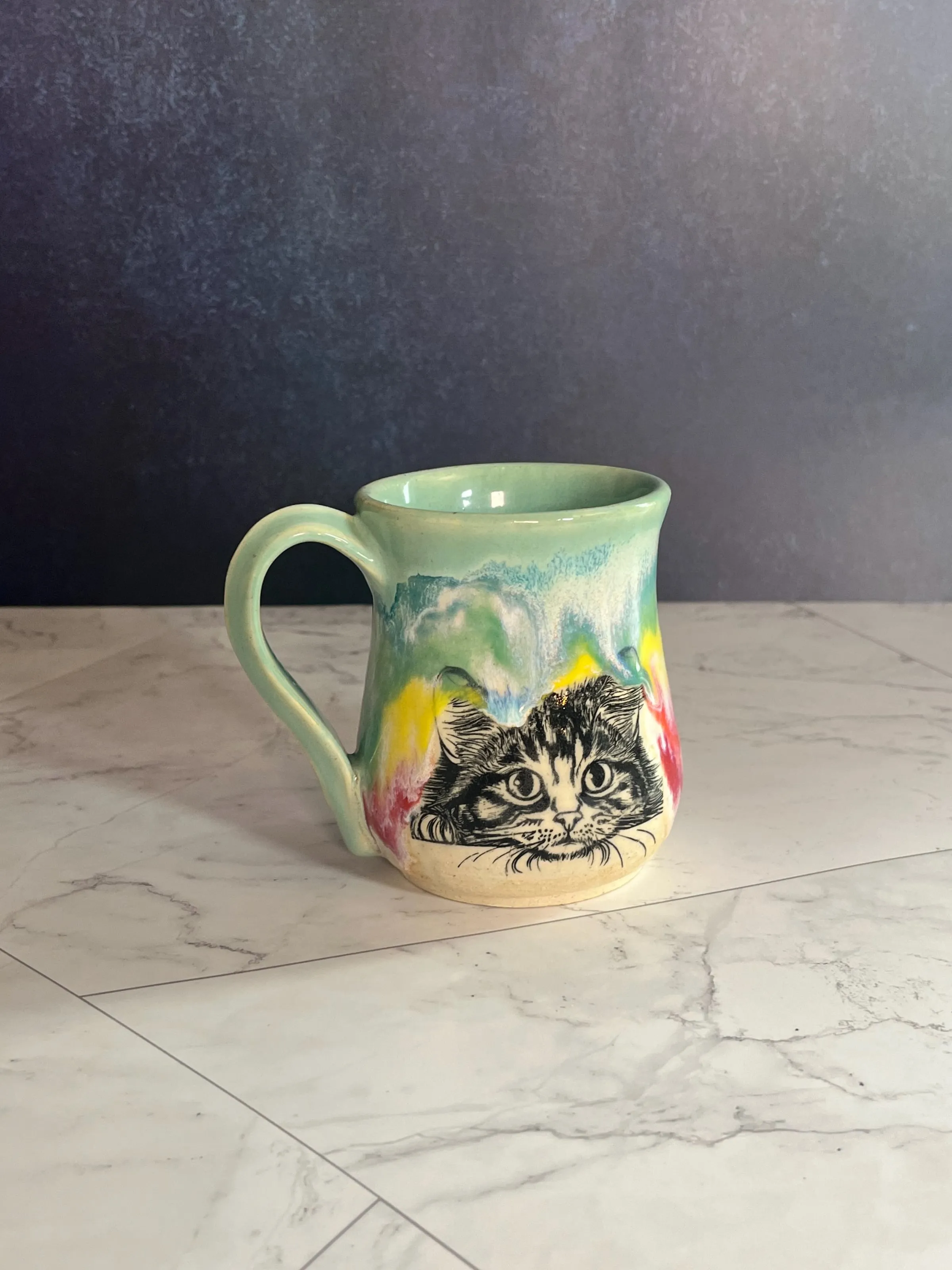 A mug with a rainbow drip glaze and a tabby cat face carved through the slip